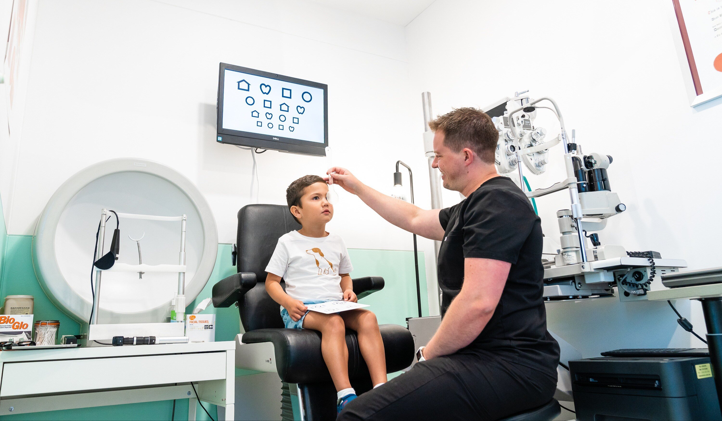 A male optometrist conducting an eye test on a young male child. there is an eye chart in the background.